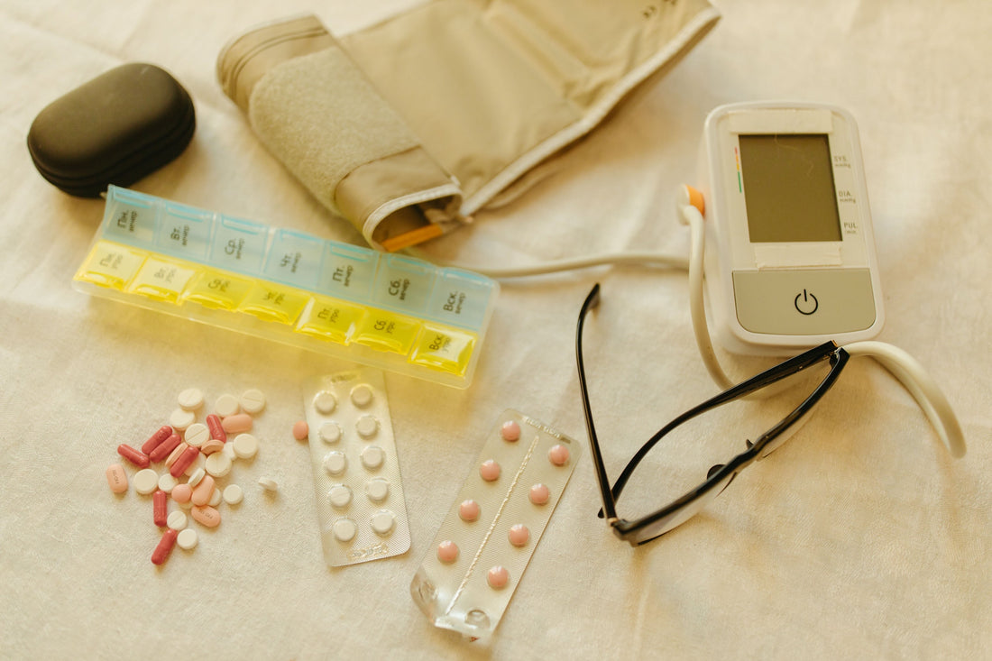 A lat lay of essential caregiving supplies including a weekly pill organizer, blood pressure monitor, medications, and glasses - blog post about some the caregiving supplies every family caregiver needs before a loved one moves in. Caregivers Collective