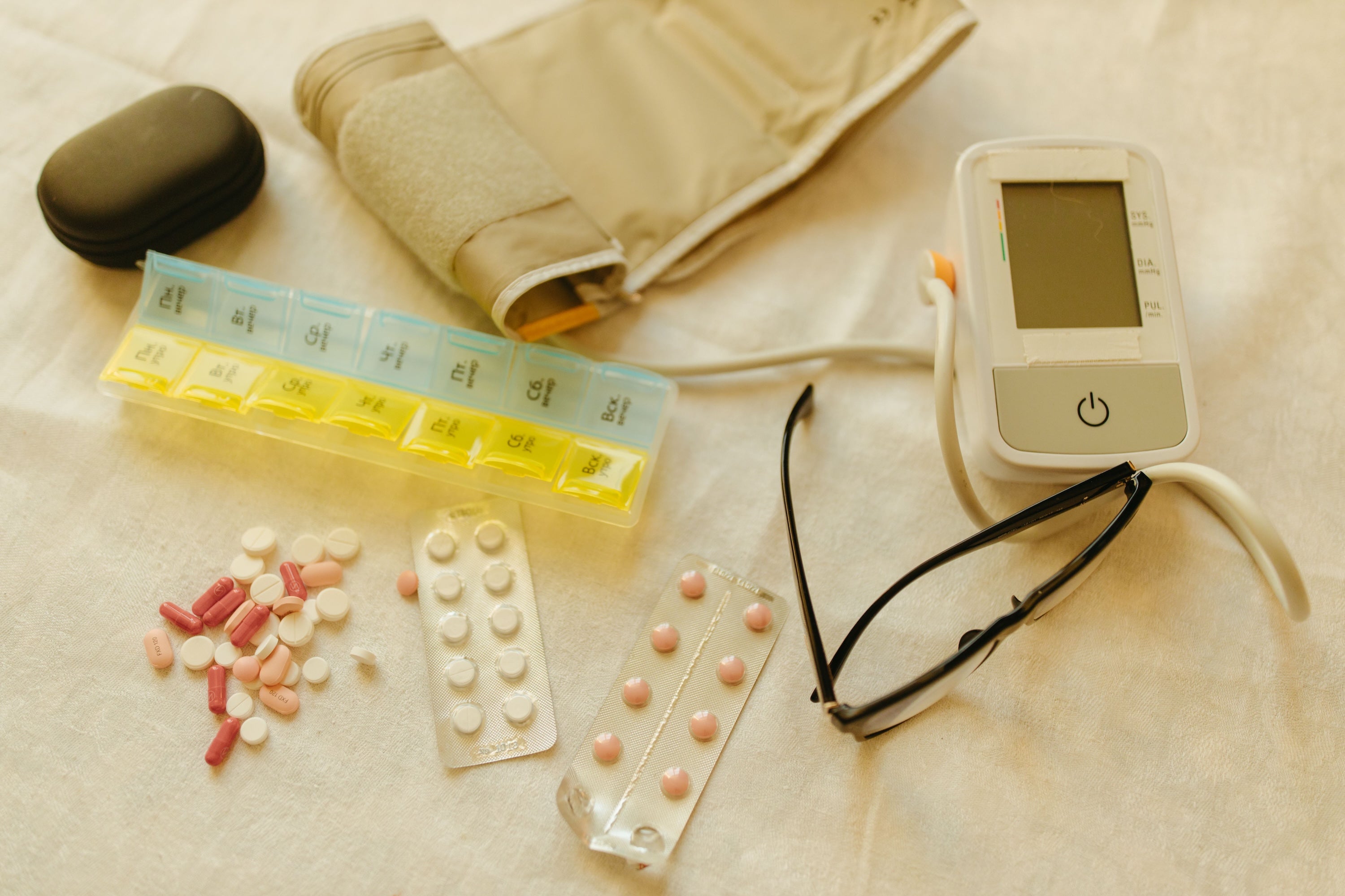 A lat lay of essential caregiving supplies including a weekly pill organizer, blood pressure monitor, medications, and glasses - blog post about some the caregiving supplies every family caregiver needs before a loved one moves in. Caregivers Collective